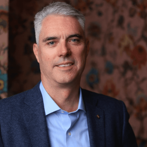 A man in a blue blazer smiling against a floral wallpaper background.